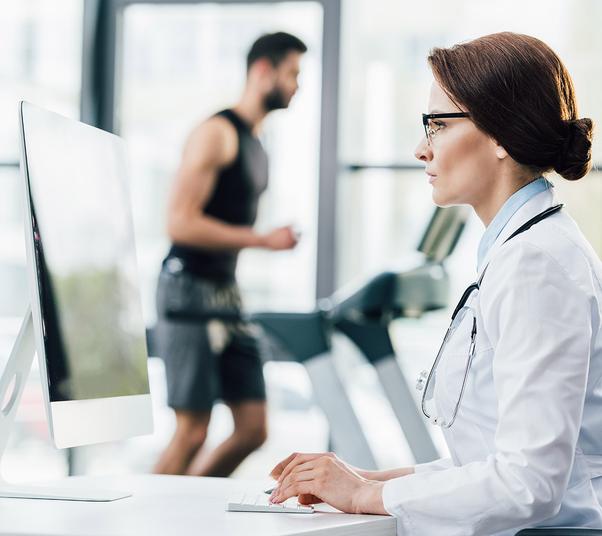 doctor in glasses using computer while sportsman running on trea